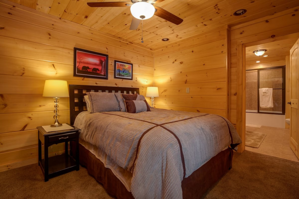 Cozy wood-paneled cabin bedroom with a large bed, two nightstands, and warm lamp light; bathroom visible through an open doorway.