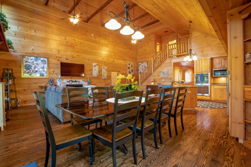 Rustic cabin interior with a long dark wood dining table and black chairs, floral centerpiece, and a staircase to a loft.