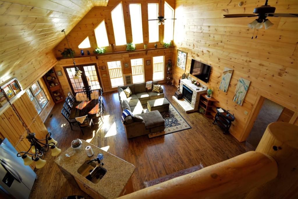 Log cabin living room with vaulted wood ceiling, tall windows, gray sectional sofa, and a stone fireplace with a mounted tv.