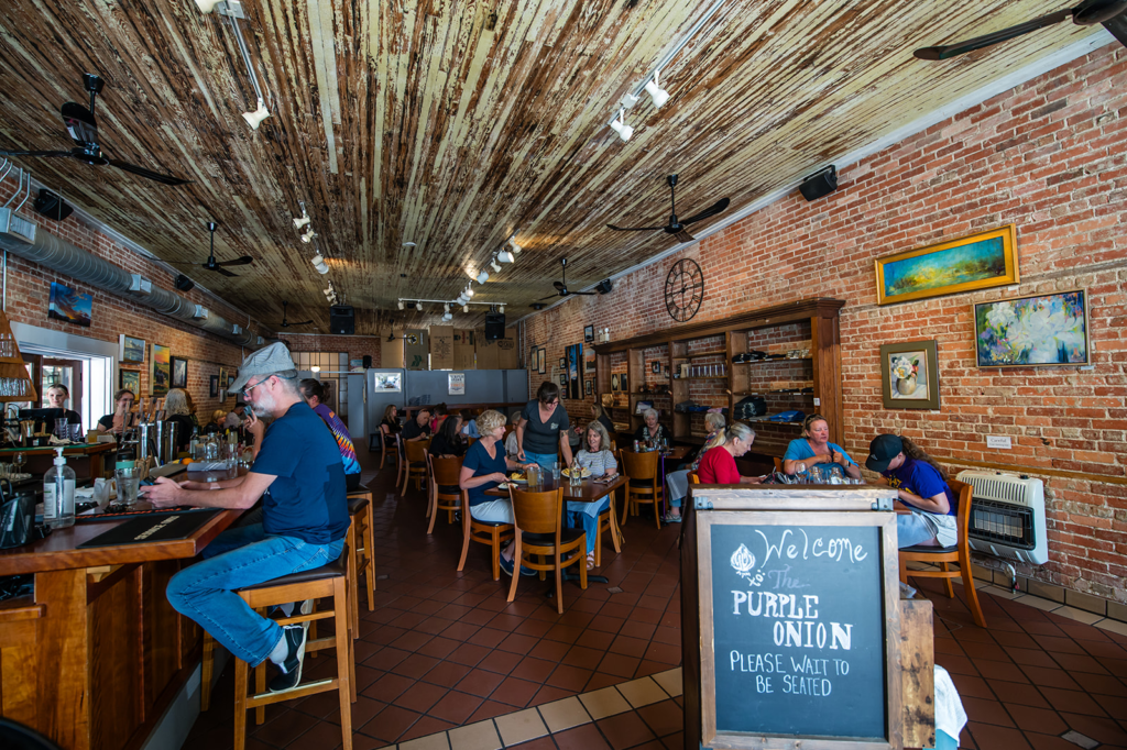 A cozy brick-walled cafe with customers seated at tables and a bar counter on the left under a weathered wooden ceiling.
