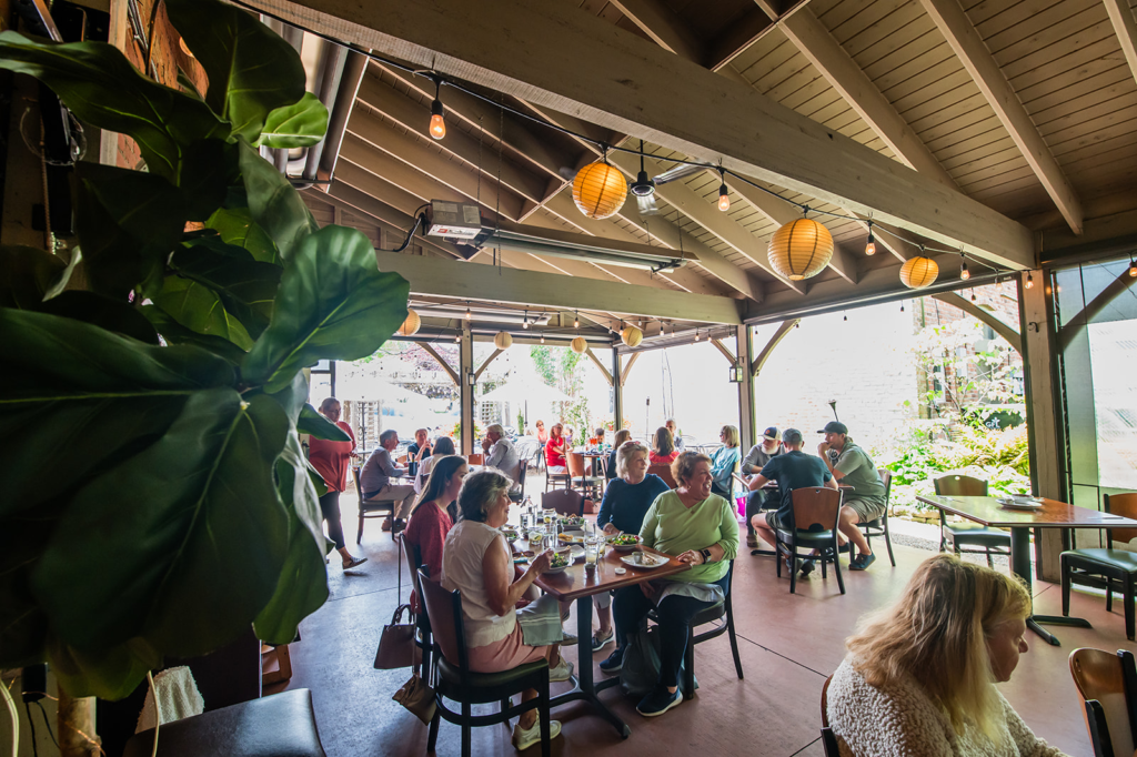 People dining at wooden tables in an open-air restaurant patio with hanging lanterns and string lights above.