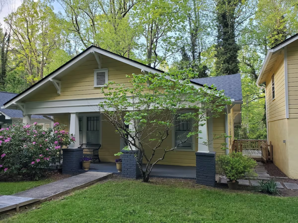 Yellow craftsman-style house with a covered front porch, porch columns, and a flowering shrub in the yard.