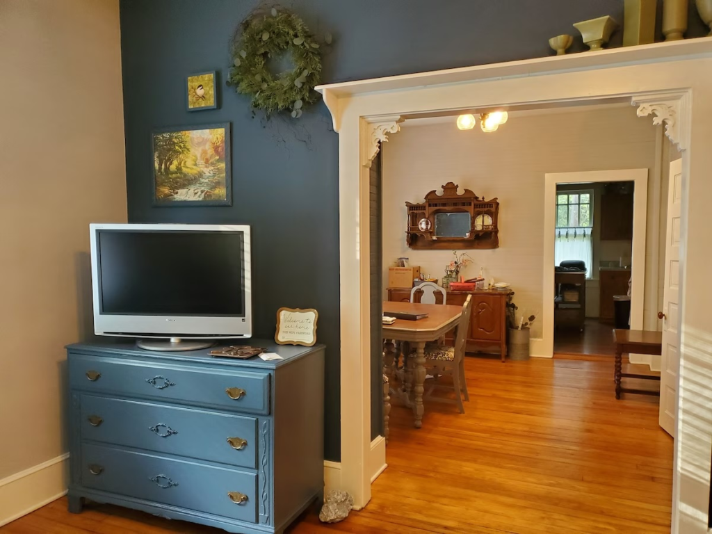 Blue dresser with a flat-screen tv on top sits against a dark blue wall, with a wreath and framed art above; doorway opens to a sunny dining room.