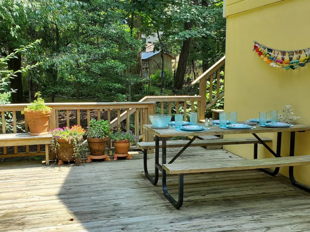 Outdoor wooden deck with a picnic table set for a meal; blue glasses and plates, potted plants along a bench, and a yellow wall nearby.