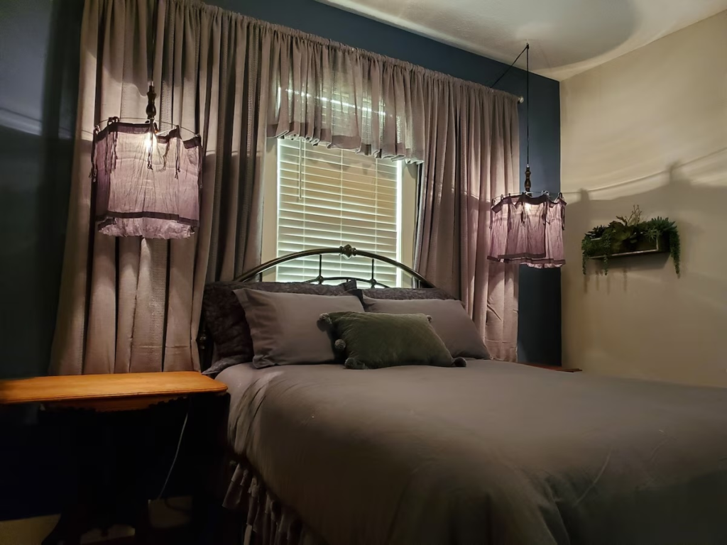 Cozy bedroom with a metal-curved bed, gray bedding, and mauve pendant lights; curtains frame a window behind the bed.