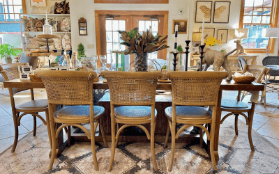 Spring Creek Interior Featured Image Long wooden dining table with wicker chairs in a bright showroom, topped with plants and decorative objects.