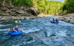 Feature Image - Tubing → Discover Saluda, NC Group of people tubing on a fast river, wearing life jackets, with rocky banks and green trees nearby.