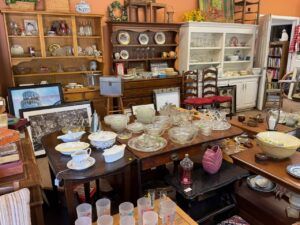 Busy antique shop interior with glass bowls, china, crystal, and framed pictures on tables and shelves.