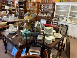 A cluttered antique shop with a dark wood table piled high with stacked china, a floral pitcher, glassware, and decorative metal lamps, shelves of porcelain and frames in the background.