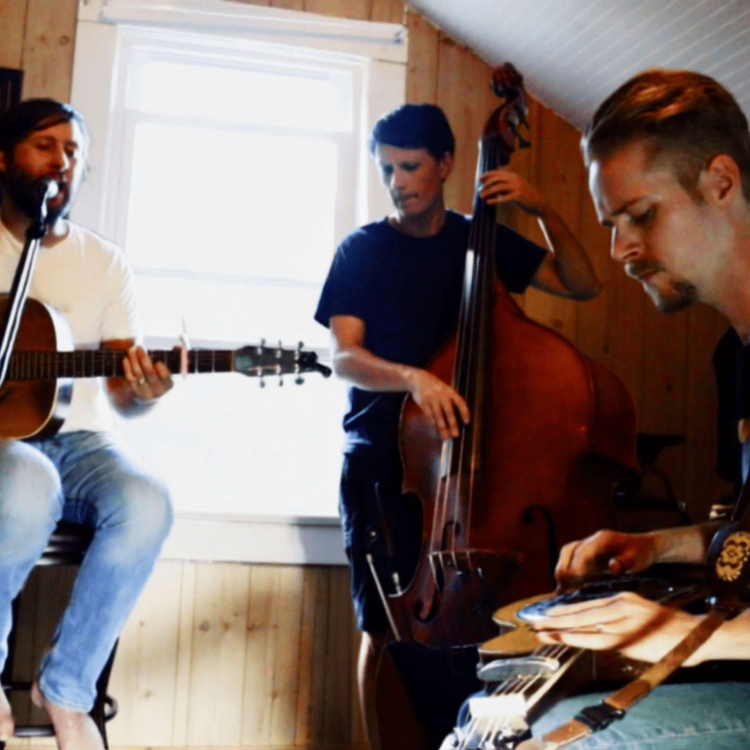 Three male musicians play guitar, upright bass, and pedal steel in a sunlit, wood-paneled room.