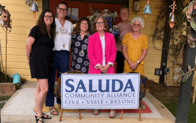 Eight people pose together on a porch behind a SALUDA Community Alliance banner reading 'LIVE • VISIT • BELONG'.