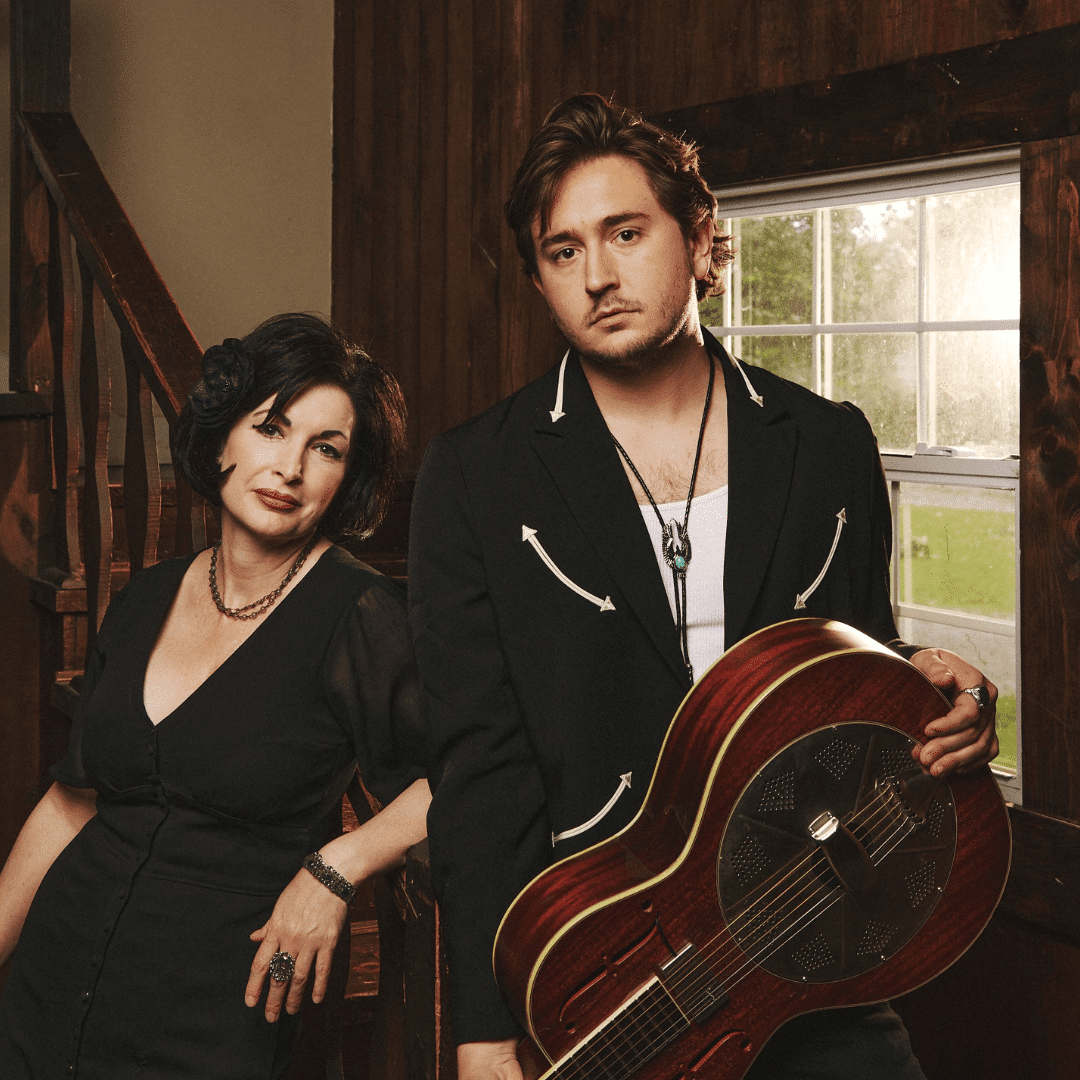 Man in a black jacket holding a wooden guitar beside a woman in a black dress in a warm, wood-paneled room by a window.
