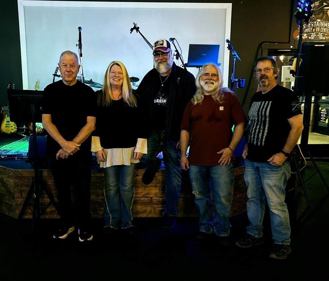 Five adults stand together on a stage in a casual group photo, with instruments and stage gear in the background.