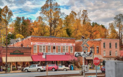 Blog - FallSaluda → Discover Saluda, NC Autumn street scene in a small town: orange-leaved trees flank brick storefronts, including Thompson's Market & Grill, with parked cars and a stop sign.
