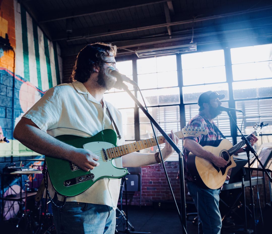 Two musicians perform with electric and acoustic guitars on a brick-walled, mural-lit stage.