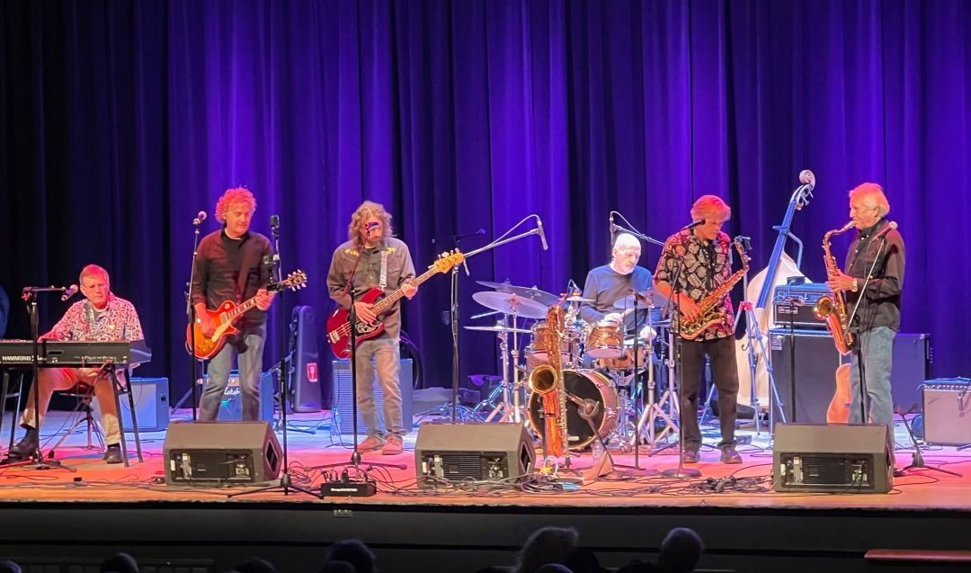 Seven-member band on stage under purple lighting; keyboardist, two guitarists, bassist, drummer, and two saxophonists performing live.
