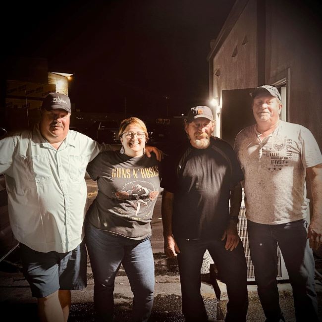 Four adults pose for a casual outdoor night photo, smiling at the camera; one wears a Guns N' Roses shirt and cap, others in casual tee shirts.