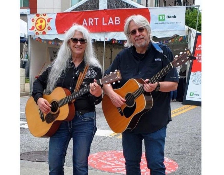 Two musicians with acoustic guitars perform at an outdoor art festival under a red 'ART LAB' banner.