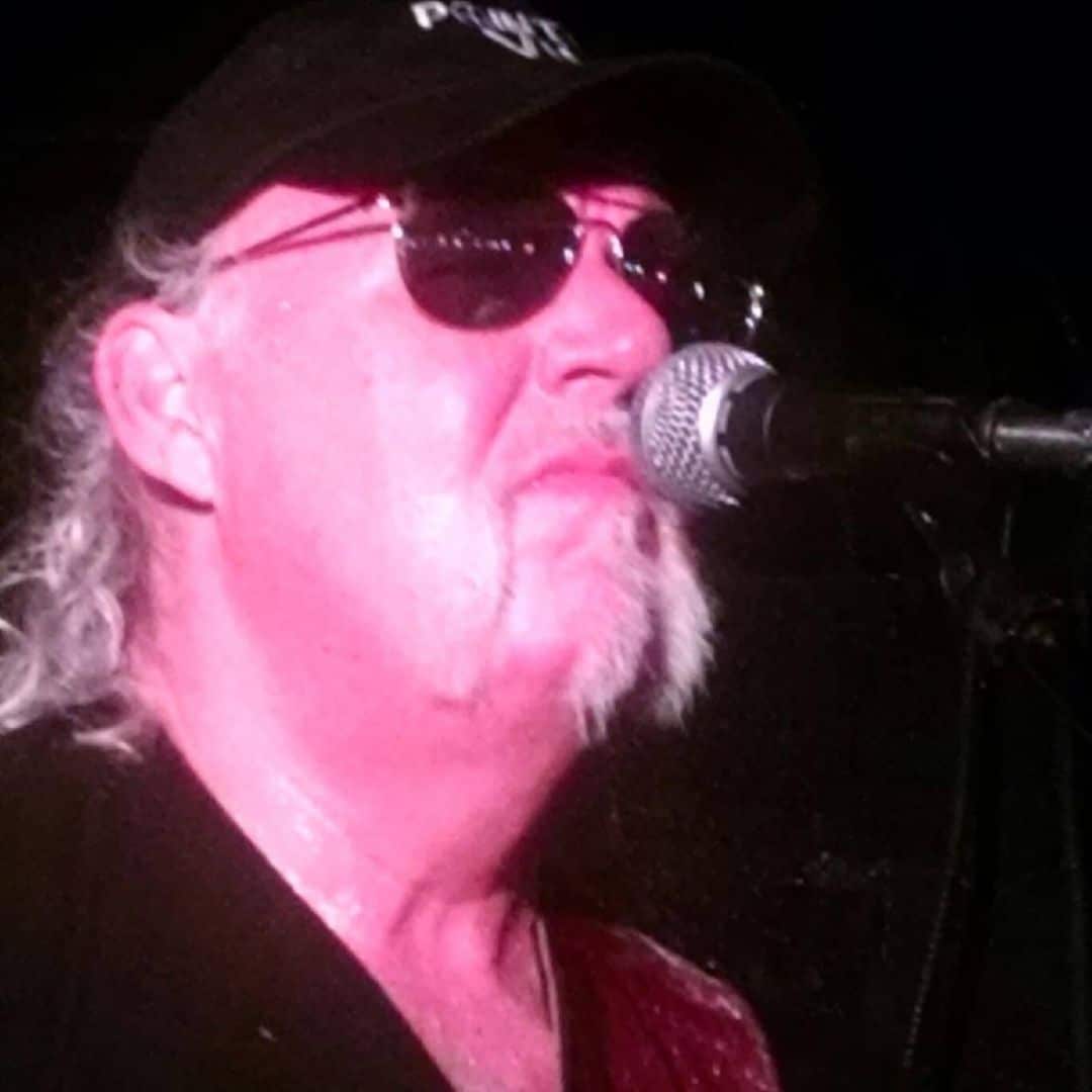 Male singer wearing a black cap and sunglasses performs into a microphone on stage under pink lighting.