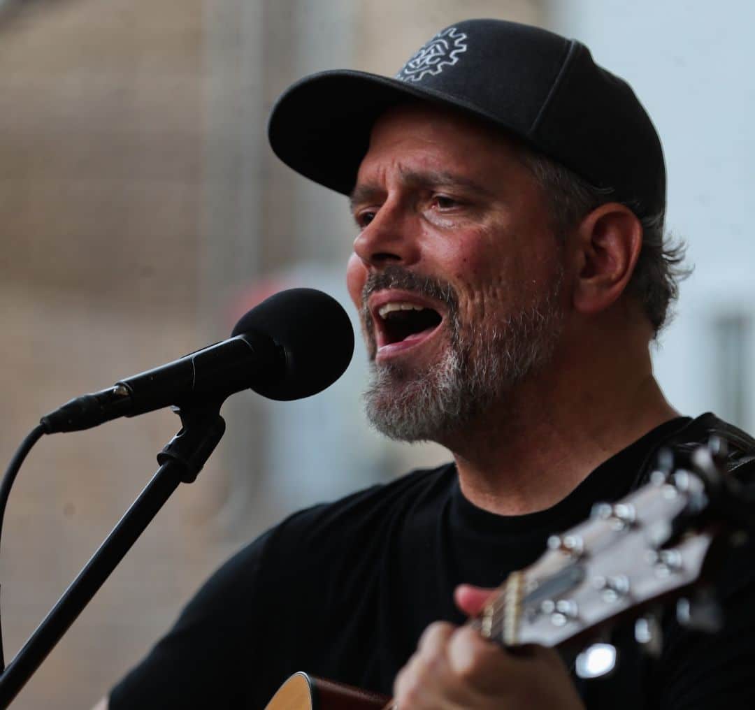 Male singer with a beard, wearing a black cap, singing into a microphone while strumming an acoustic guitar.