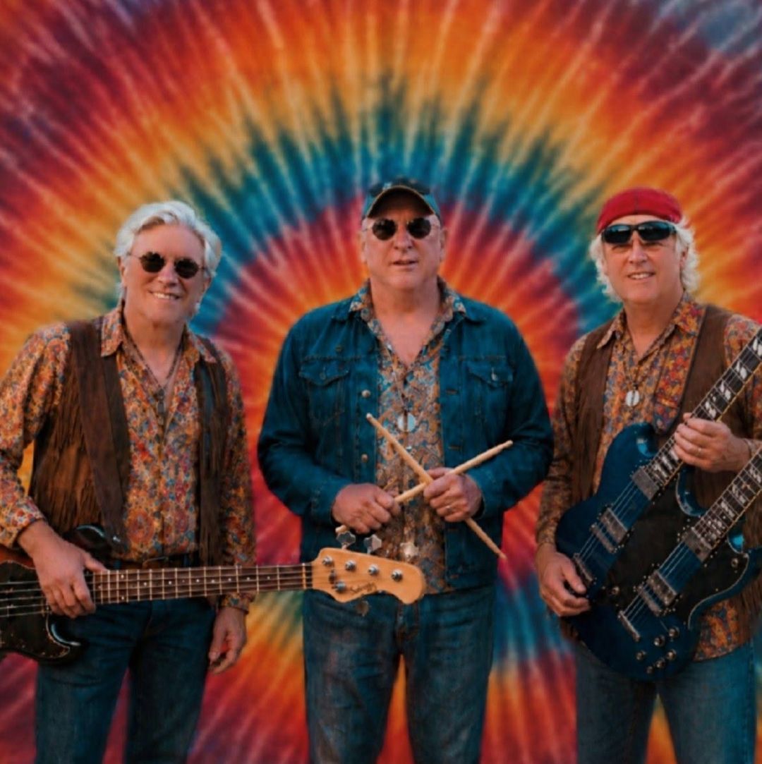 Three musicians pose with guitars in front of a vibrant tie-dye backdrop.