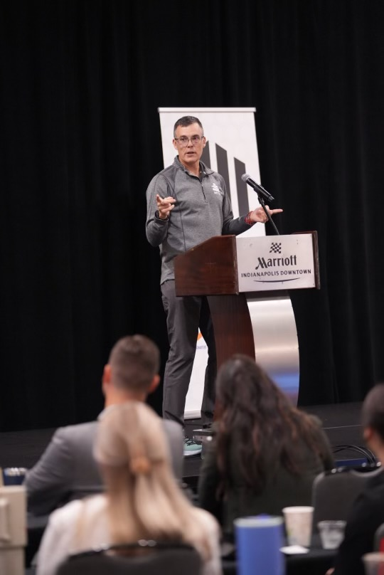 Speaker at a podium with a microphone, Marriott Indianapolis Downtown sign visible, addressing an audience in a conference room.