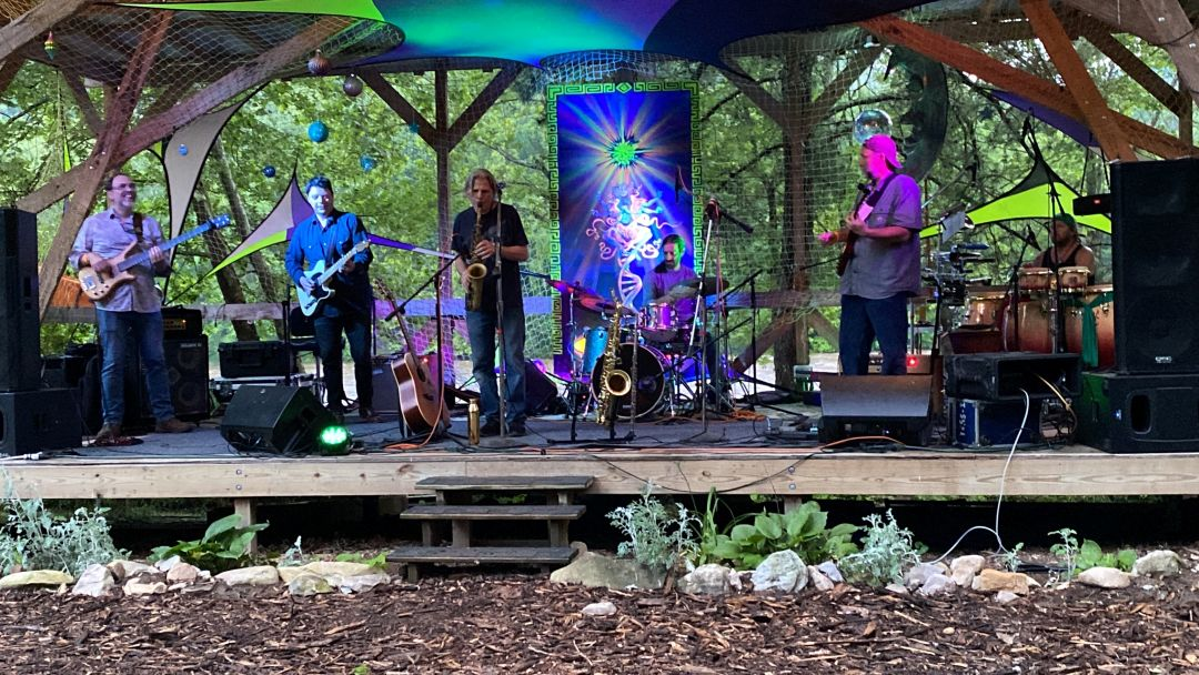 Six-piece band performing on an outdoor wooden stage surrounded by trees, with guitars, saxophones, and drums in a forest setting behind a colorful banner.