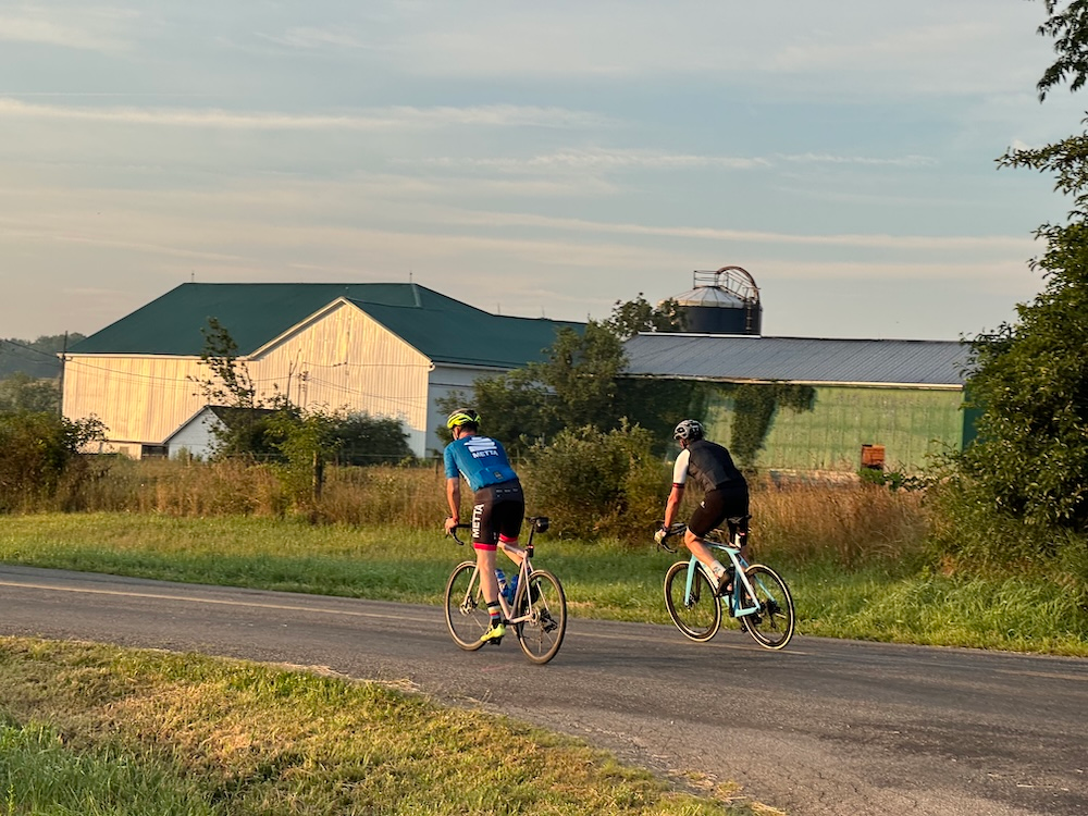 Two cyclists ride along a rural road with a large green-roofed barn and fields in the background.