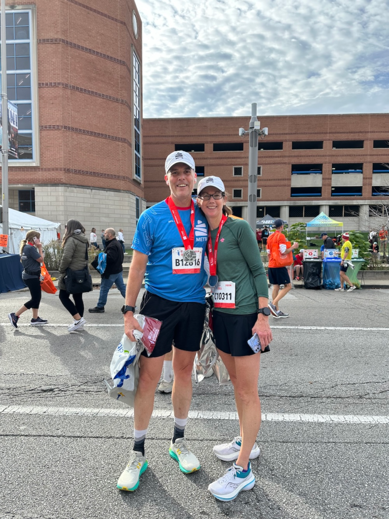 Two runners with medals posing together after a race in an urban finish area, smiling at the camera under a blue sky.