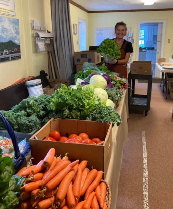 Woman in an apron holds a head of lettuce behind a long table of fresh vegetables, including carrots, kale, cabbage, and tomatoes, in an indoor market room.