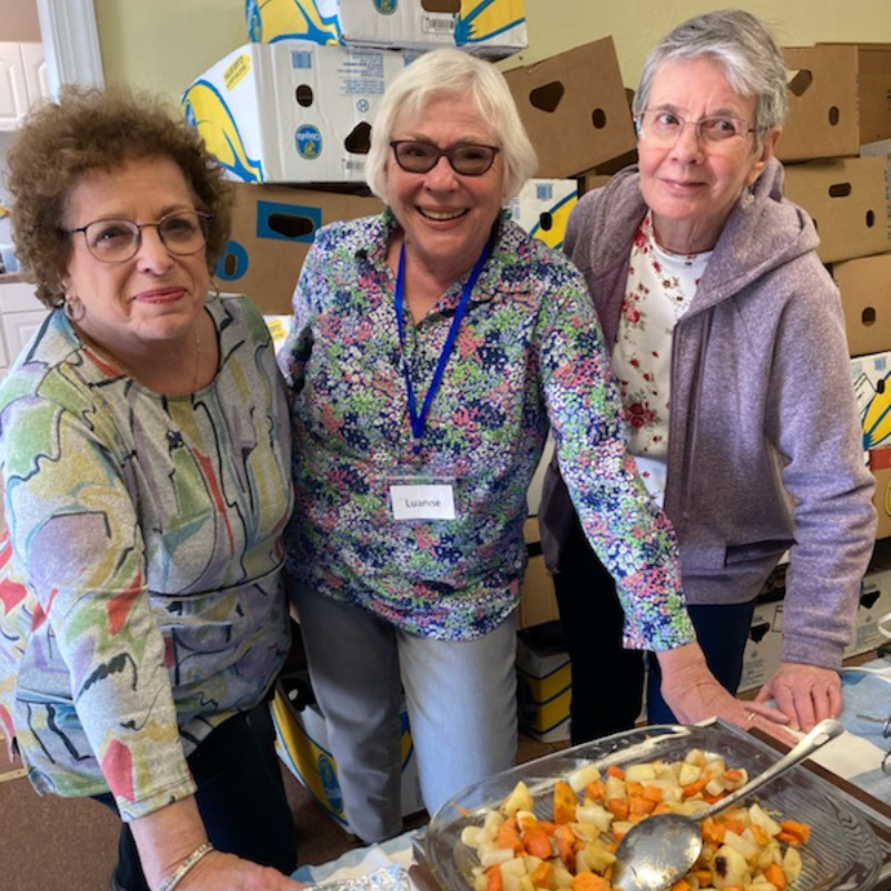 Three smiling women behind a table of fruit salad, with cardboard boxes in the background; nametag reads Luanne.