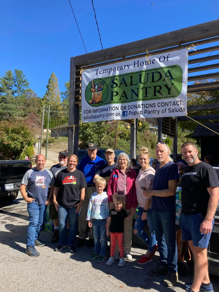 Group of people posing for a photo outdoors beneath a banner that reads 'Temporary Home Of Saluda Pantry'; clear blue sky and trees in the background, some holding bags and smiling at the camera.