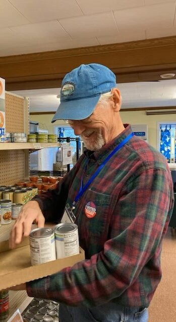 Man in a blue cap and plaid shirt stocks canned goods in a grocery store, smiling as he places cans into a cardboard holder.