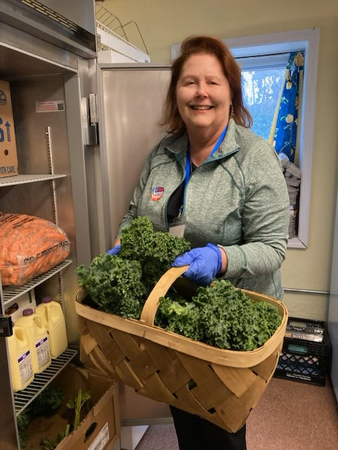 Smiling woman in a gray jacket and blue gloves holding a wooden basket filled with kale near a fridge in a kitchen or food prep area.
