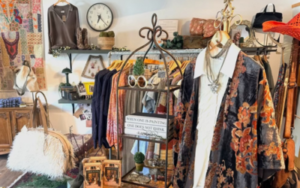 Boho boutique interior with clothing racks, a floral kimono on a mannequin, wicker baskets, plants, shelves, and a clock on the wall.