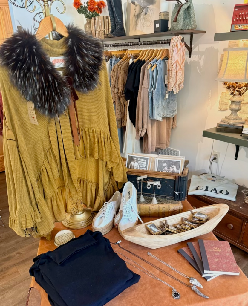 A mustard yellow dress with a large dark fur collar on a hanger in a boutique display, surrounded by shelves and clothing racks.