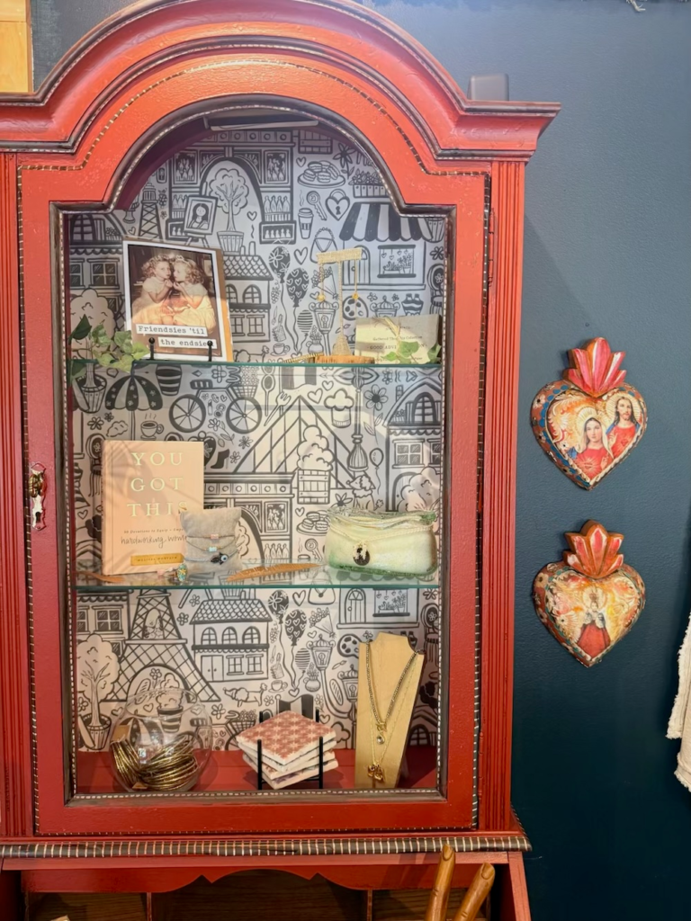 Red display cabinet with a black-and-white patterned interior, holding a framed photo, a 'YOU GOT THIS' book, jewelry display, and decorative hearts on blue wall.