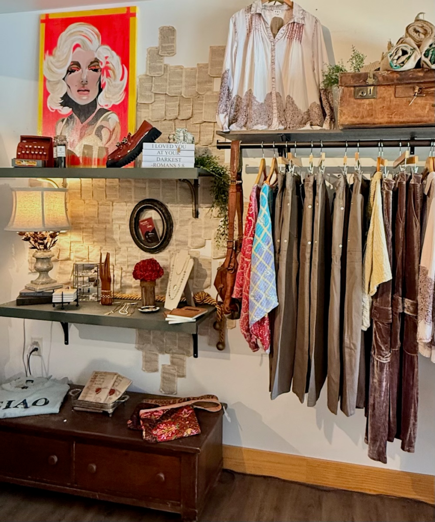 Boutique display with a rack of neutral-toned pants and dresses, colorful scarves, and a white embroidered blouse on top shelf beside vintage props and a wooden trunk.