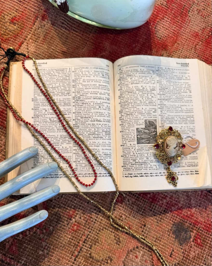 Open dictionary with gold and red bead necklaces draped across pages, and an ornate cameo brooch on the right side.