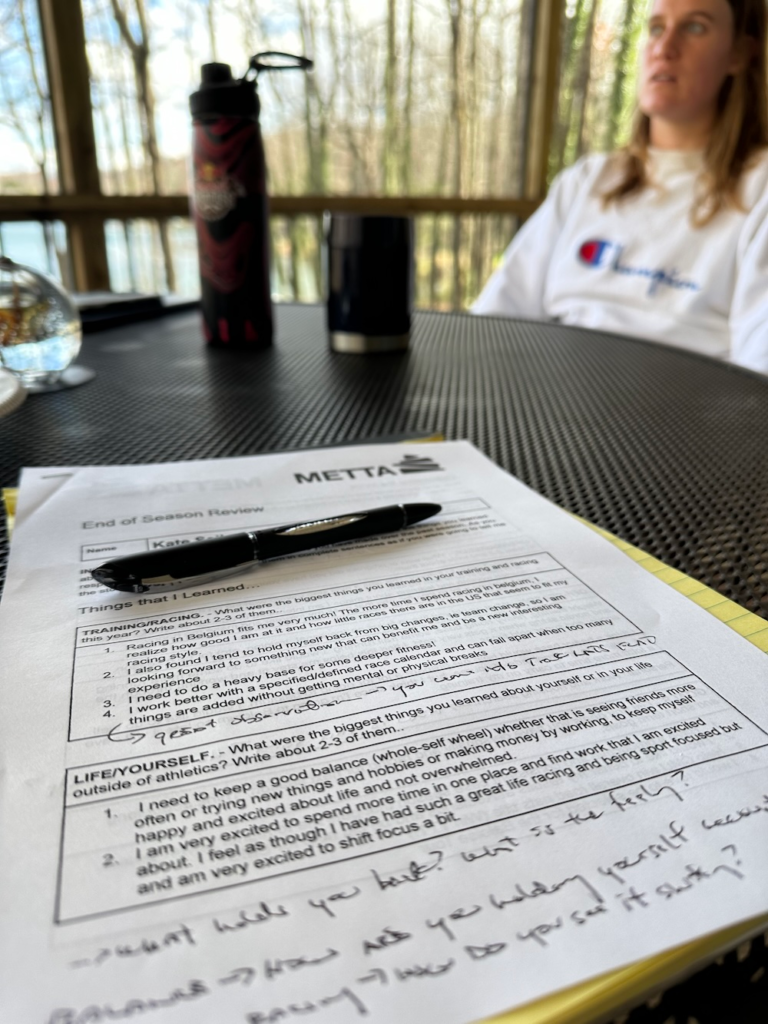 Close-up of a printed worksheet titled 'End of Season Review' on a black mesh table, with a black pen across it, a water bottle nearby, and a blurred person in a white Champion shirt in the background on a sunny outdoor deck.