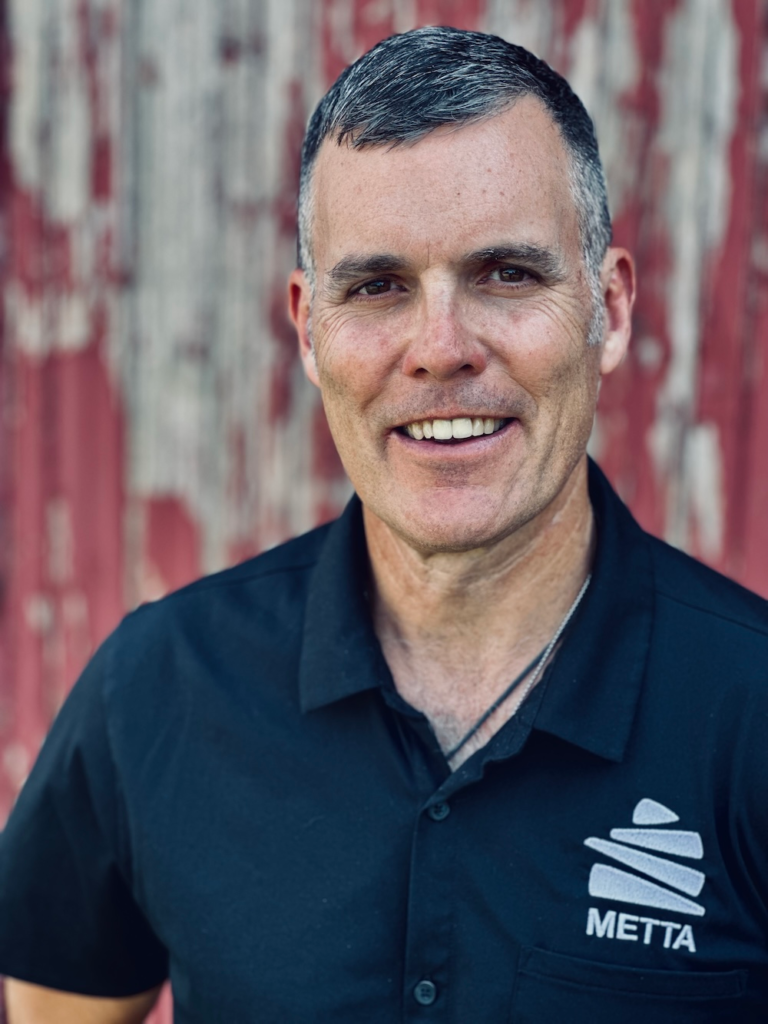 Smiling man with short gray hair in a black polo with METTA logo, standing in front of a weathered red barn.